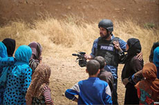 A man wearing a press vest, interviewing a group of children and women